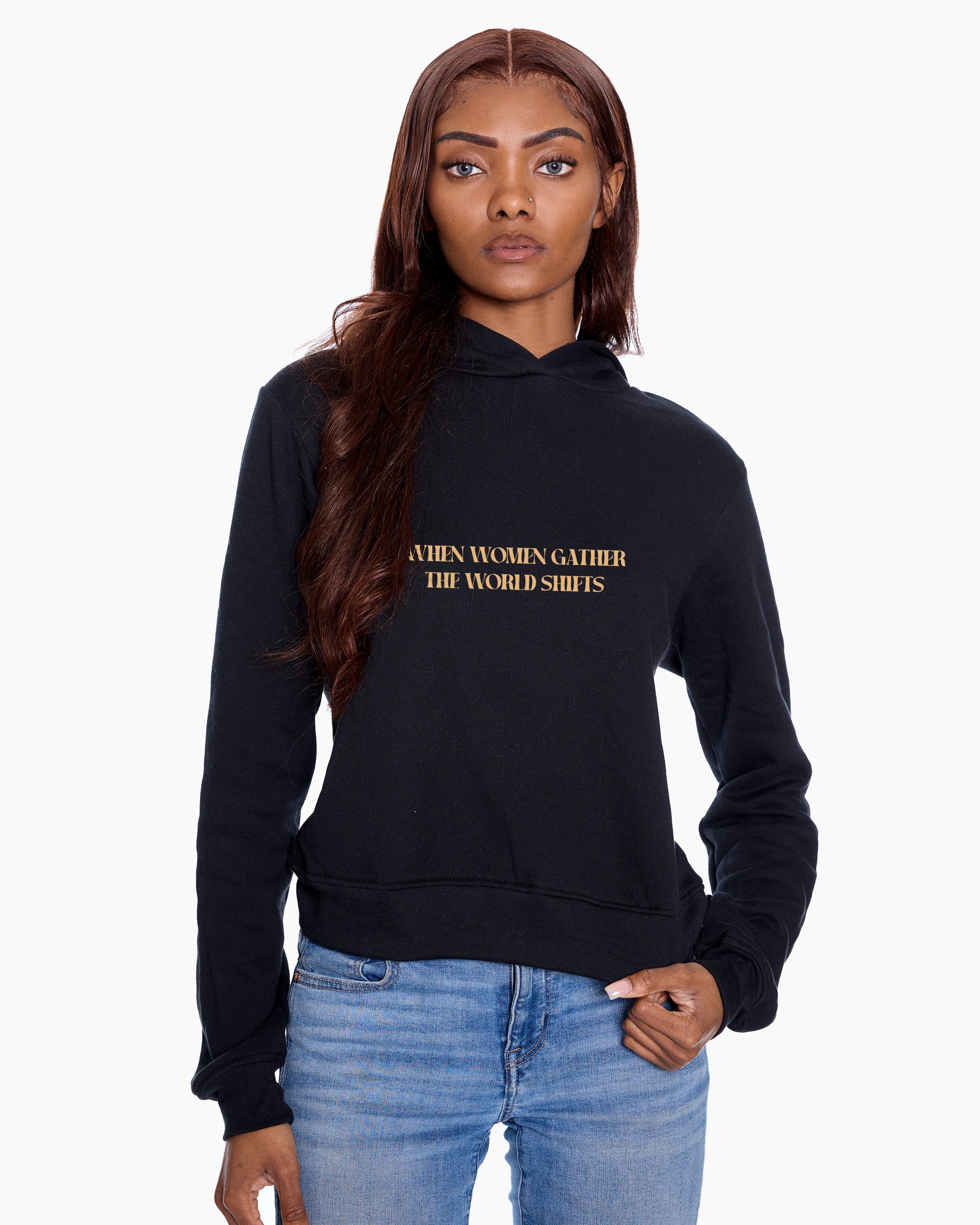 Woman wearing a black hoodie with "When Women Gather The World Shifts" graphic, standing against a white background