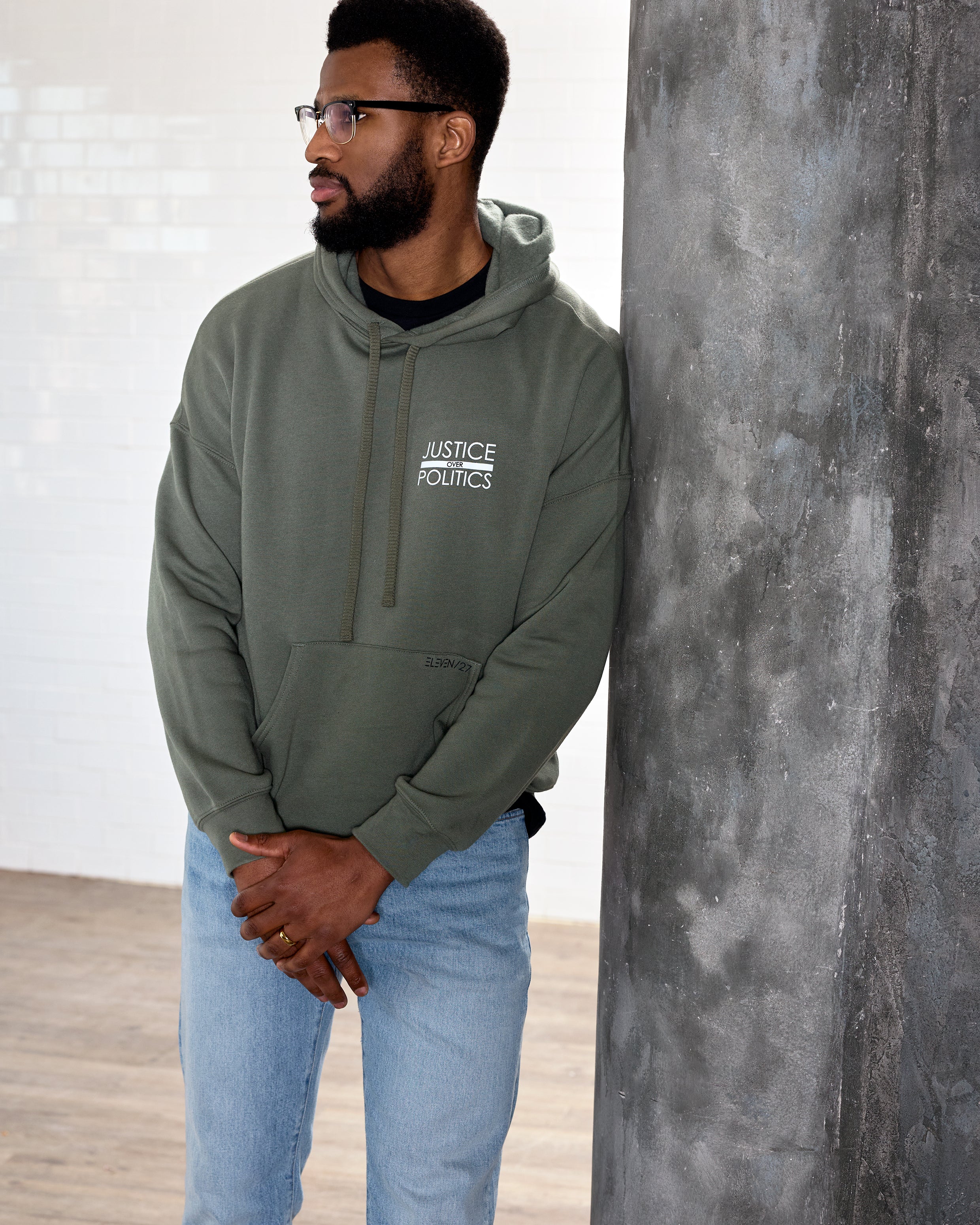 A male model wearing a green full-length hoodie with a "Justice Over Politics" graphic on the front and a logo on the back, featuring a front kangaroo pocket and drop shoulder design.