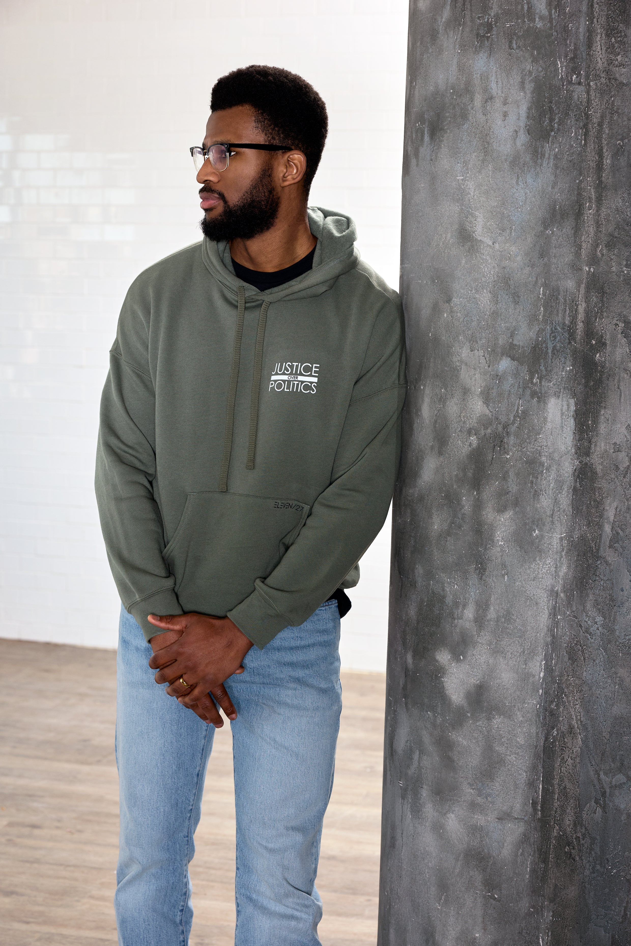 A male model wearing a green full-length hoodie with a "Justice Over Politics" graphic on the front and a logo on the back, featuring a front kangaroo pocket and drop shoulder design.