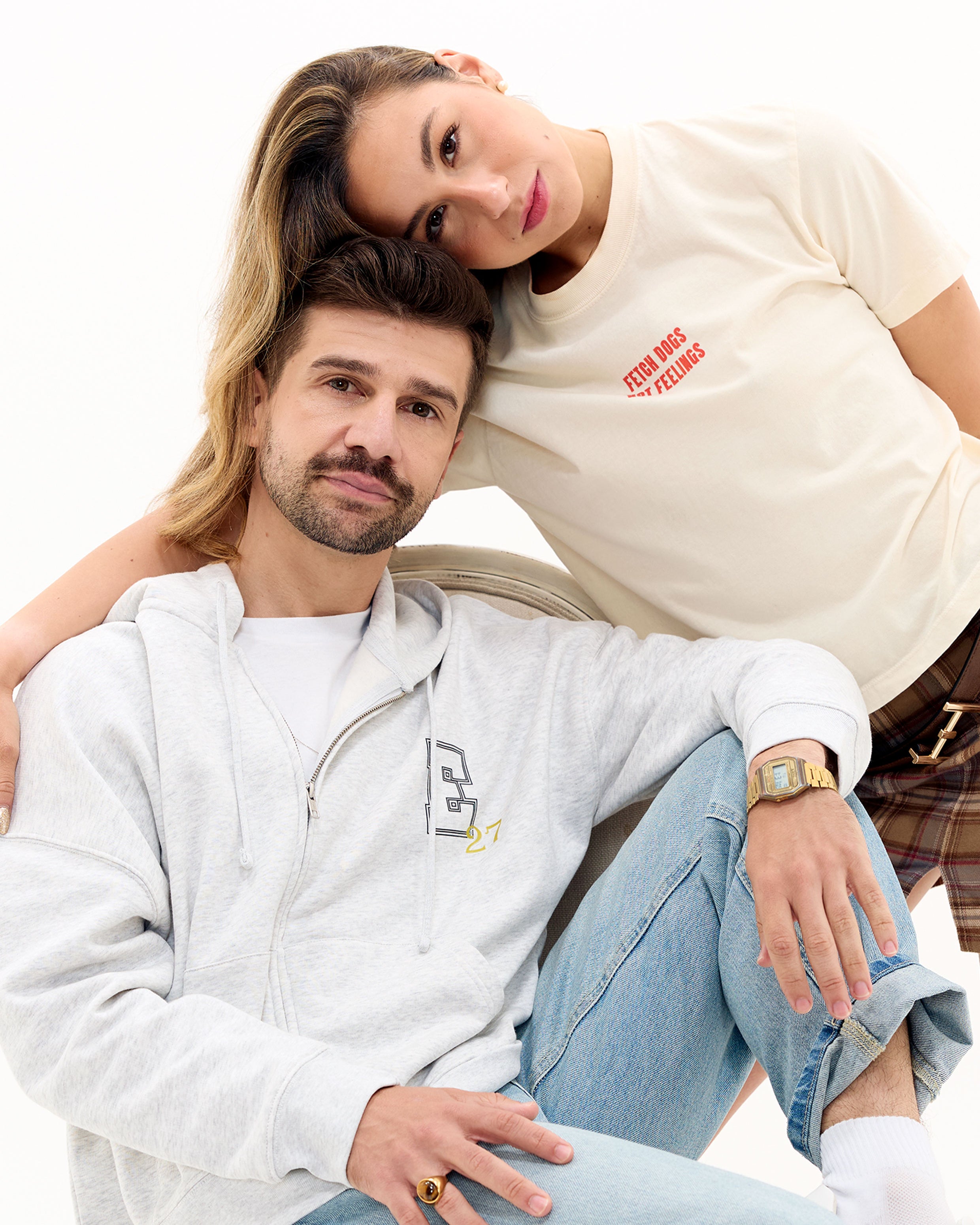 Two people sitting close together, one wearing a grey zip hoodie with an E27 logo and the other in an off-white shirt with Fetch Dogs Not Feelings graphic on the front.