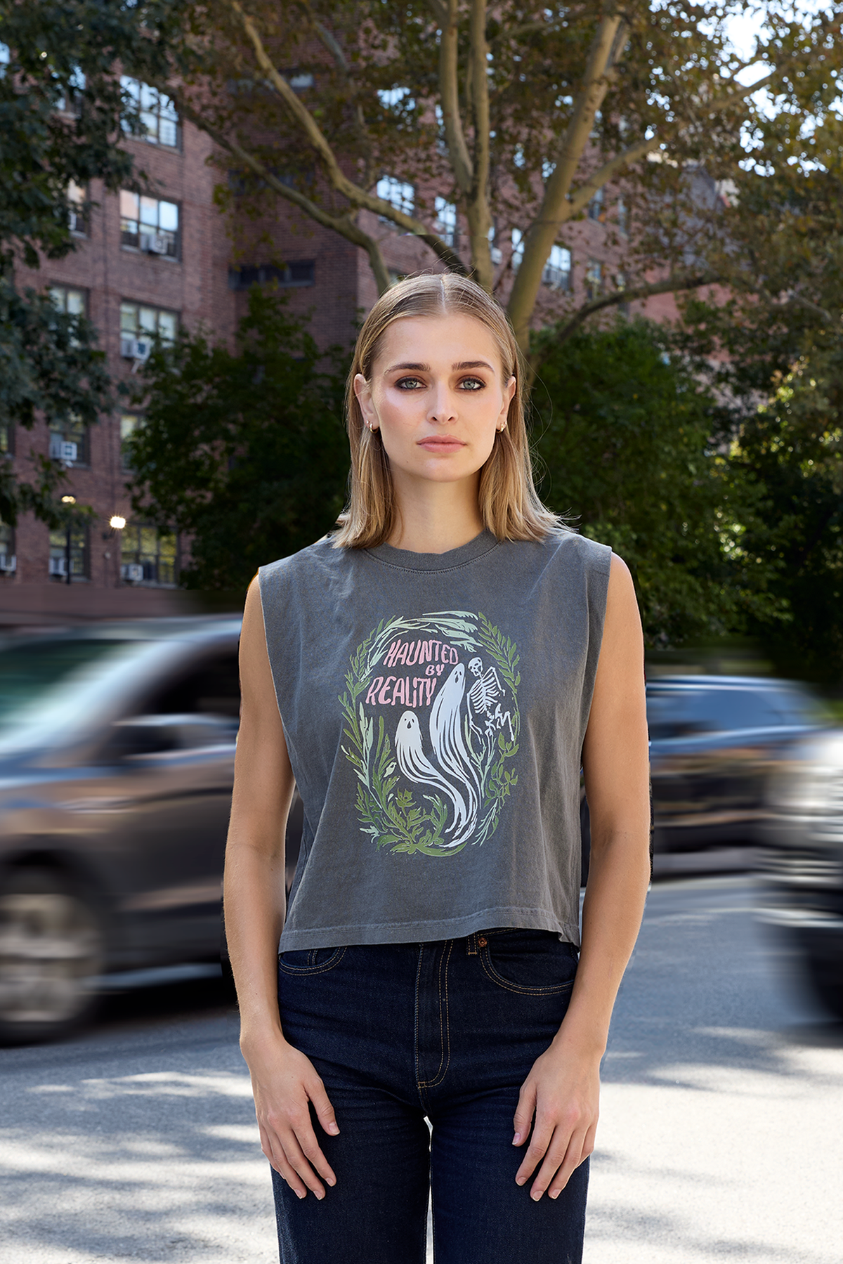 A woman stands outdoors on a city street wearing a gray sleeveless cropped graphic T-shirt with a white and green ghost illustration and the words 'Haunted By Reality"