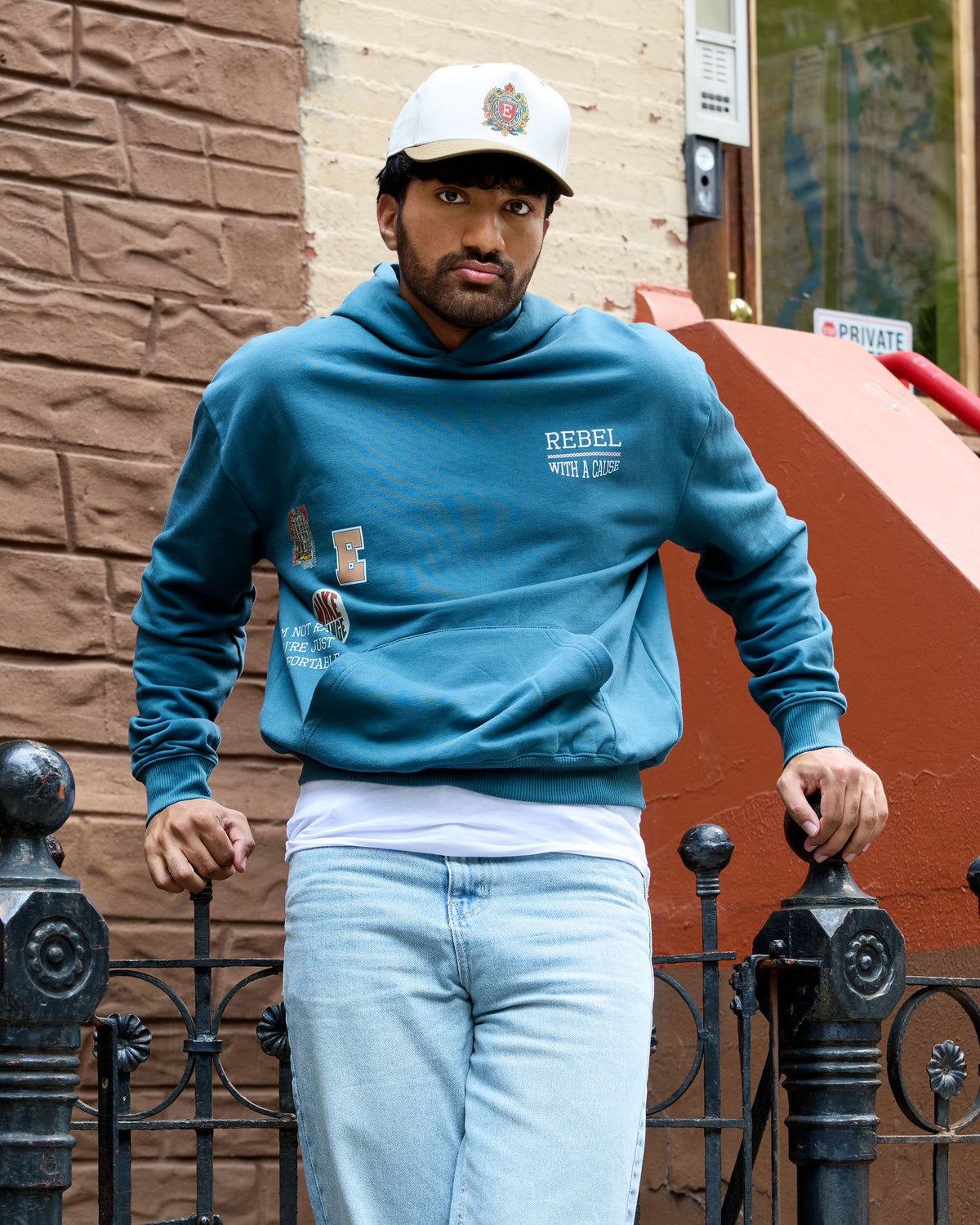 Man wearing a teal Eleven27 sweatshirt with 'Rebel With A Cause' text and light blue jeans standing against a brick wall.