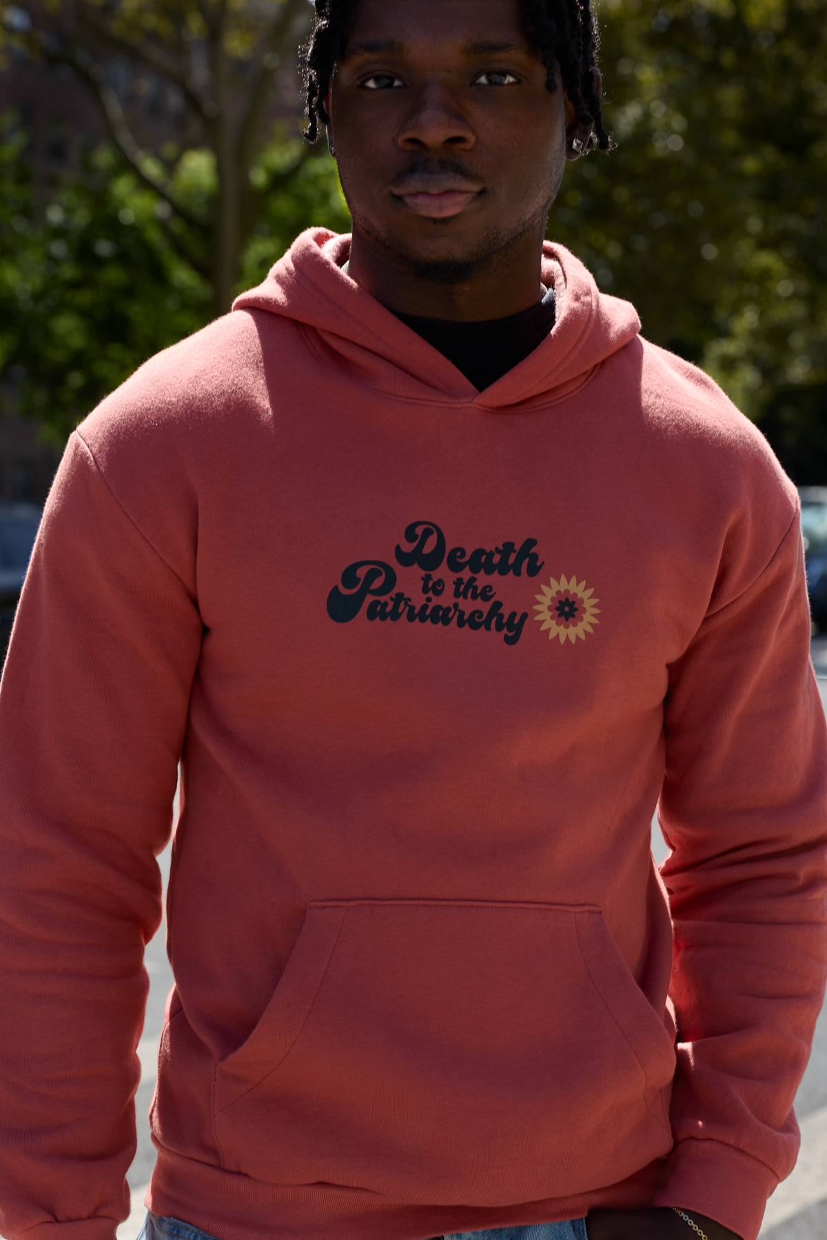 A man wearing a coral pink hoodie with the text 'Death to the Patriarchy' and a small sunflower graphic on the chest, photographed outdoors with trees in the background.