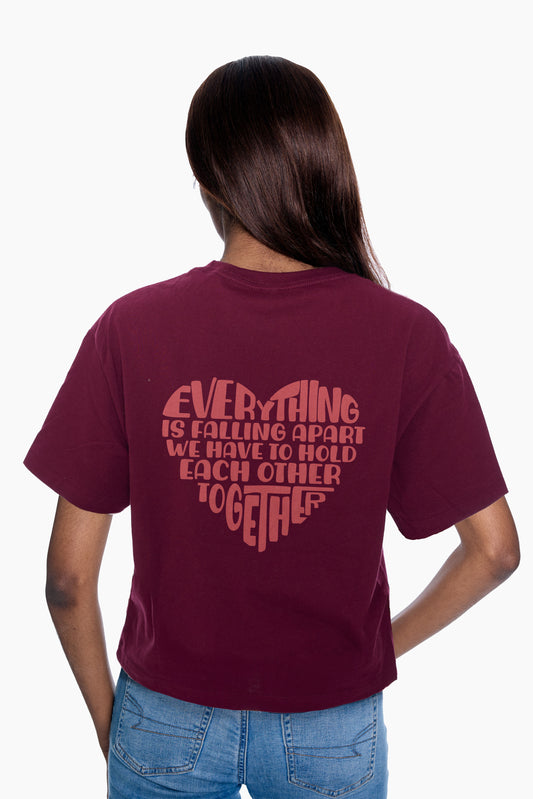 Person wearing a burgundy t-shirt with a "Everything is falling apart, we have to hold each other together" heart graphic on the back, standing against a white background.