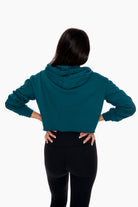 Back view of a woman in a teal cropped hoodie, hands on hips, black leggings