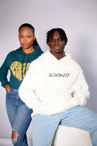 black female model with a teal cropped hoodie with a eleven27 gold heart logo on the front next to a black male model with an ivory hoodie with an eleven27 silicon logo on the front