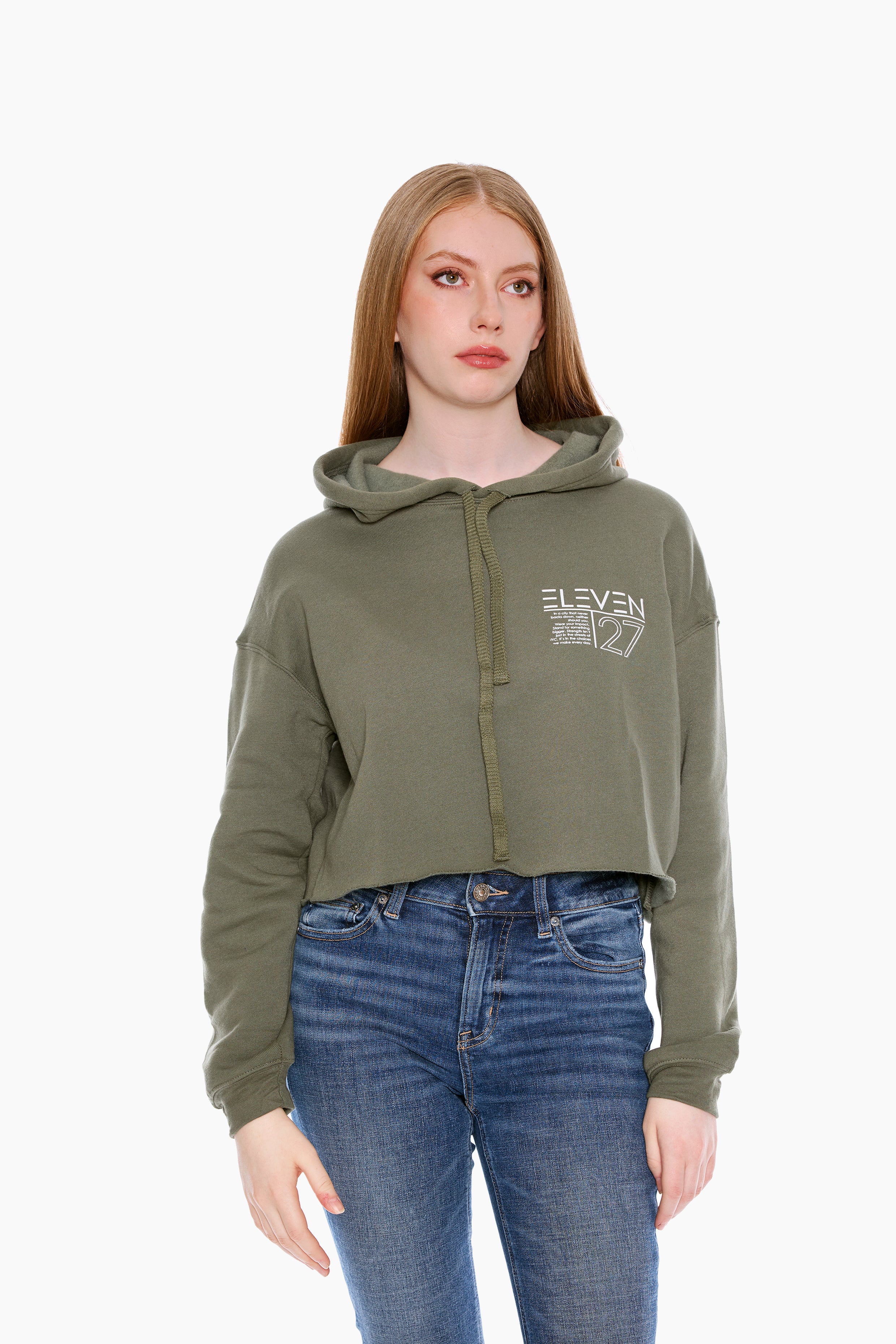 A female model wearing a green Nexus crop hoodie with a hood, drop shoulder raw cut hem, and a front logo. She is also wearing blue jeans.