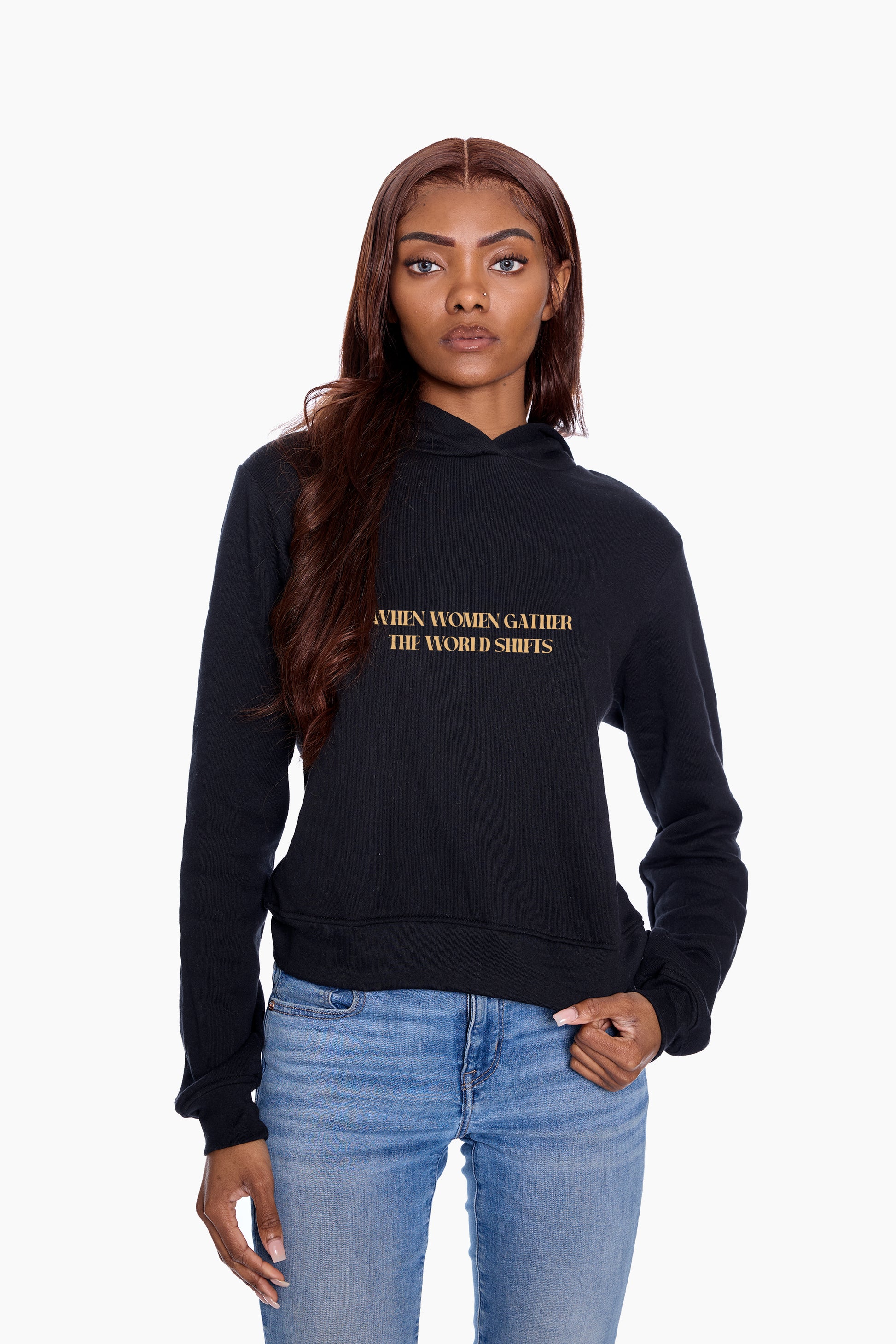 Woman wearing a black hoodie with "When Women Gather The World Shifts" graphic, standing against a white background
