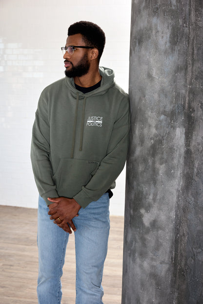 A male model wearing a green full-length hoodie with a "Justice Over Politics" graphic on the front and a logo on the back, featuring a front kangaroo pocket and drop shoulder design.
