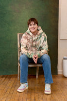 Person wearing a tie dye hoodie sitting on a chair against a green wall. Eleven27