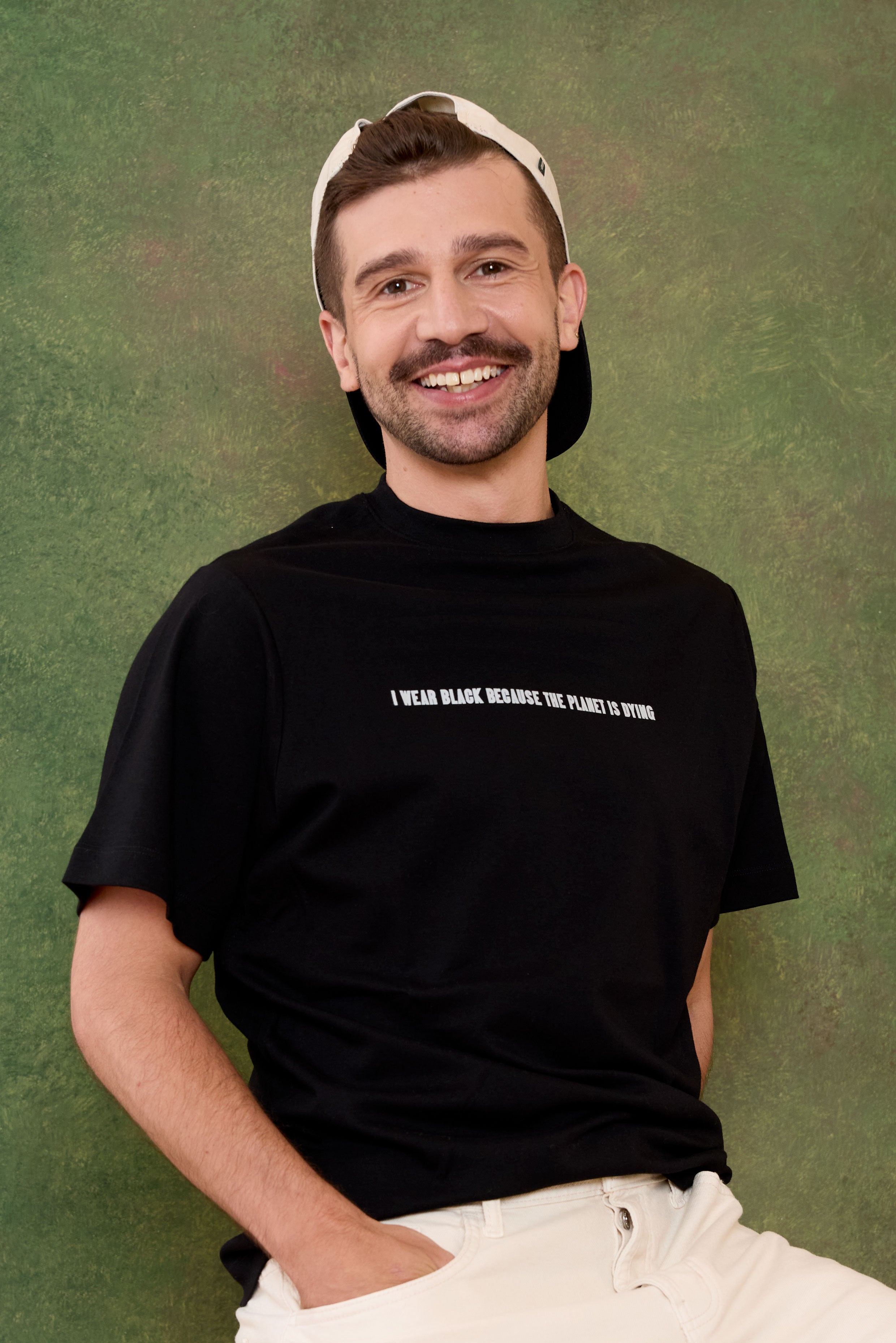Man wearing a black t-shirt with "I wear black because the planet is dying" text and a cap, sitting against a green background. Eleven27