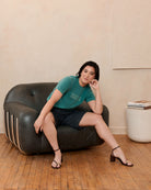 Woman wearing a green shirt with "Lipgloss & Liberation" graphic on the front, sitting on a black leather chair in a room with wooden flooring. Eleven27.