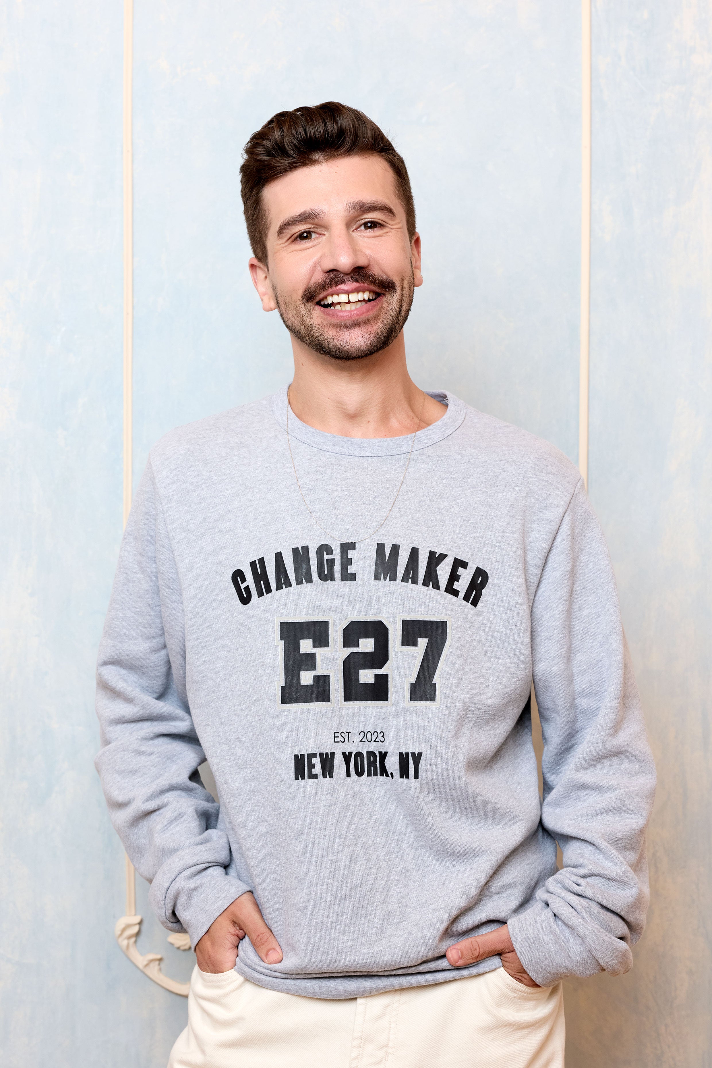 Man wearing a gray sweatshirt with 'CHANGE MAKER E27' text, standing against a light blue wall.