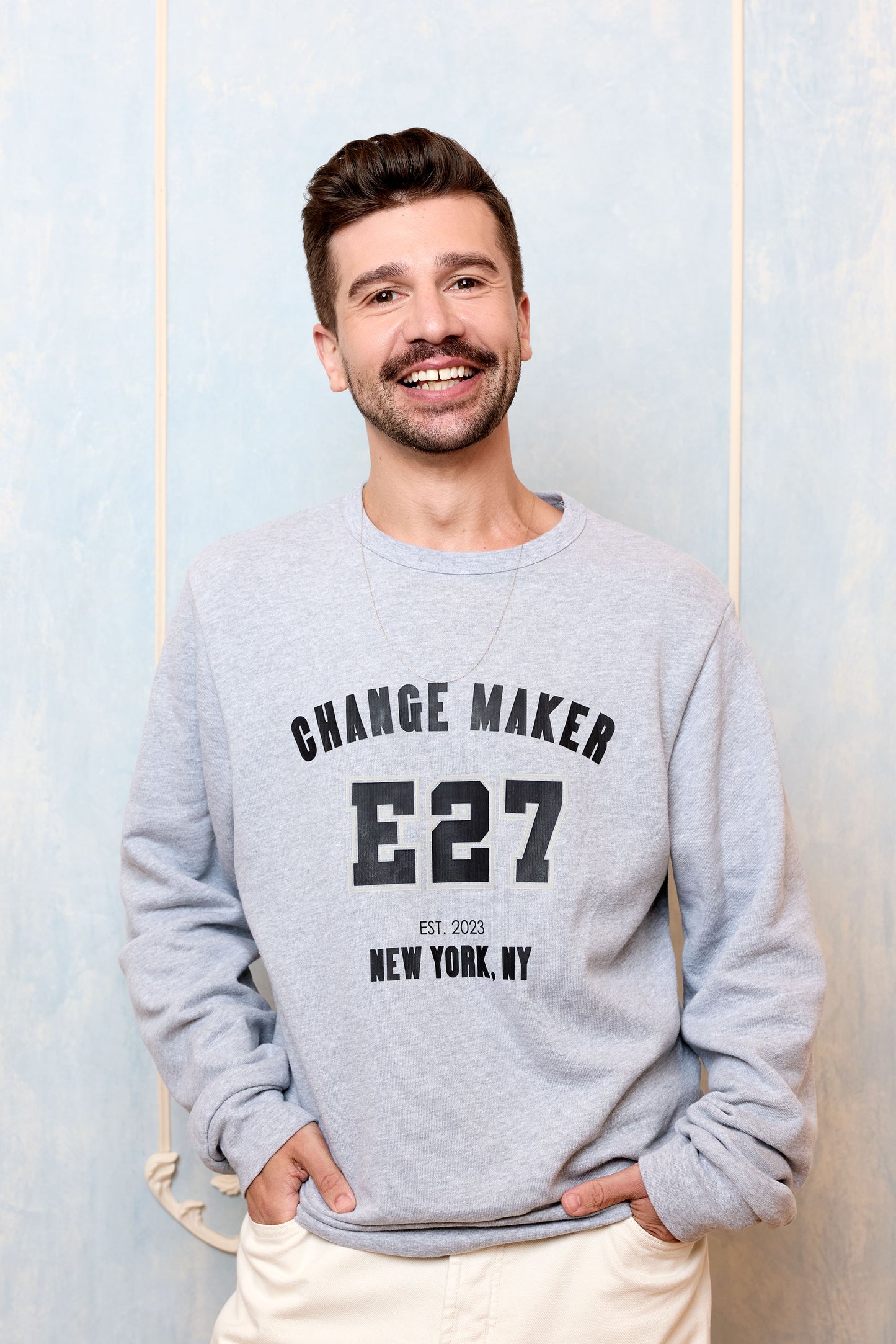 Man wearing a gray sweatshirt with 'CHANGE MAKER E27' text, standing against a light blue wall.