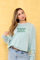 Woman wearing a light blue seafoam sweatshirt with "Oppression Ruins My Aesthetic" text on the front, against a yellow background. Eleven27