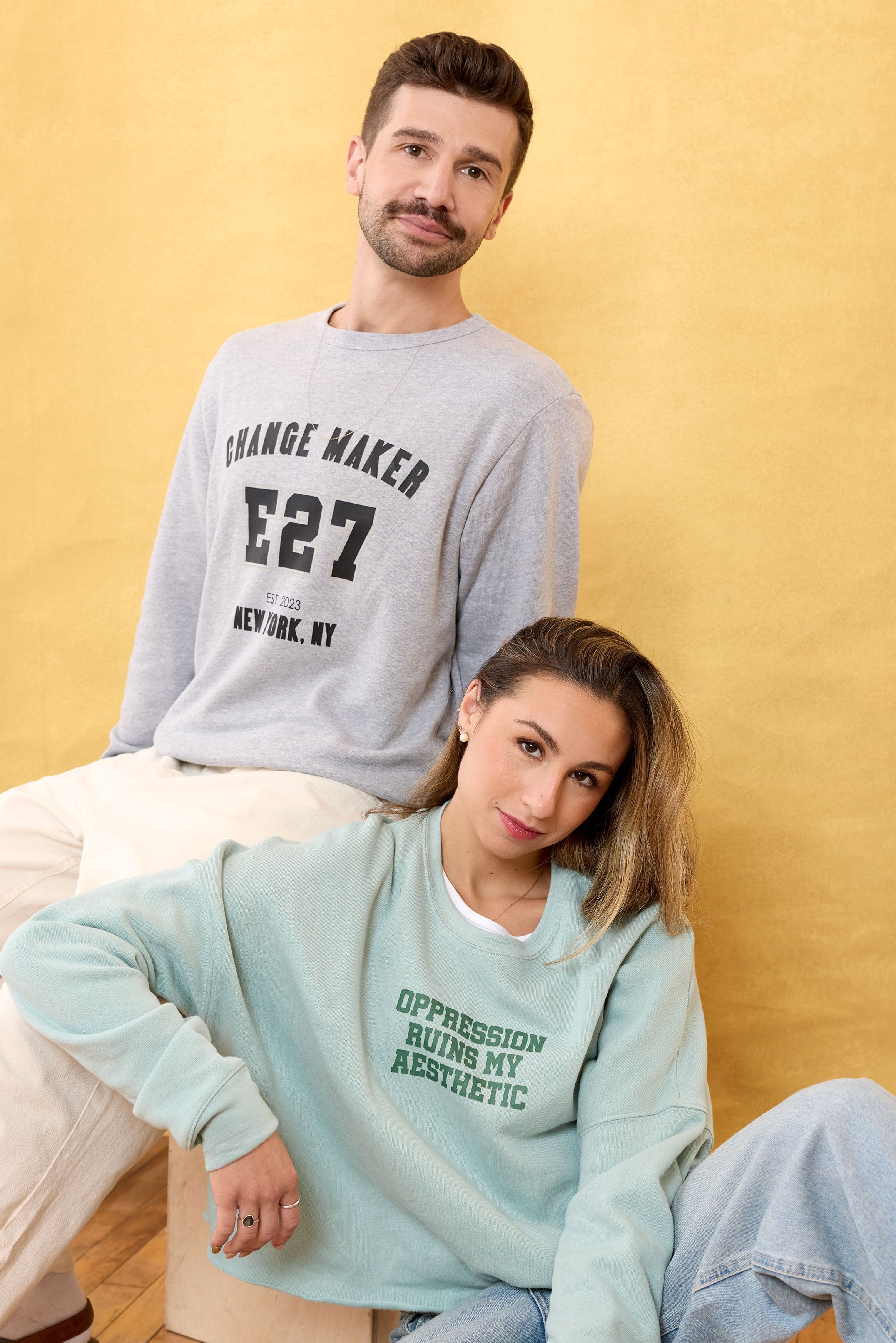 Two people wearing gray and light blue sweatshirts with text against a yellow background. Eleven27. Changemaker sweatshirt. Oppression Ruins My Aesthetic Sweatshirt
