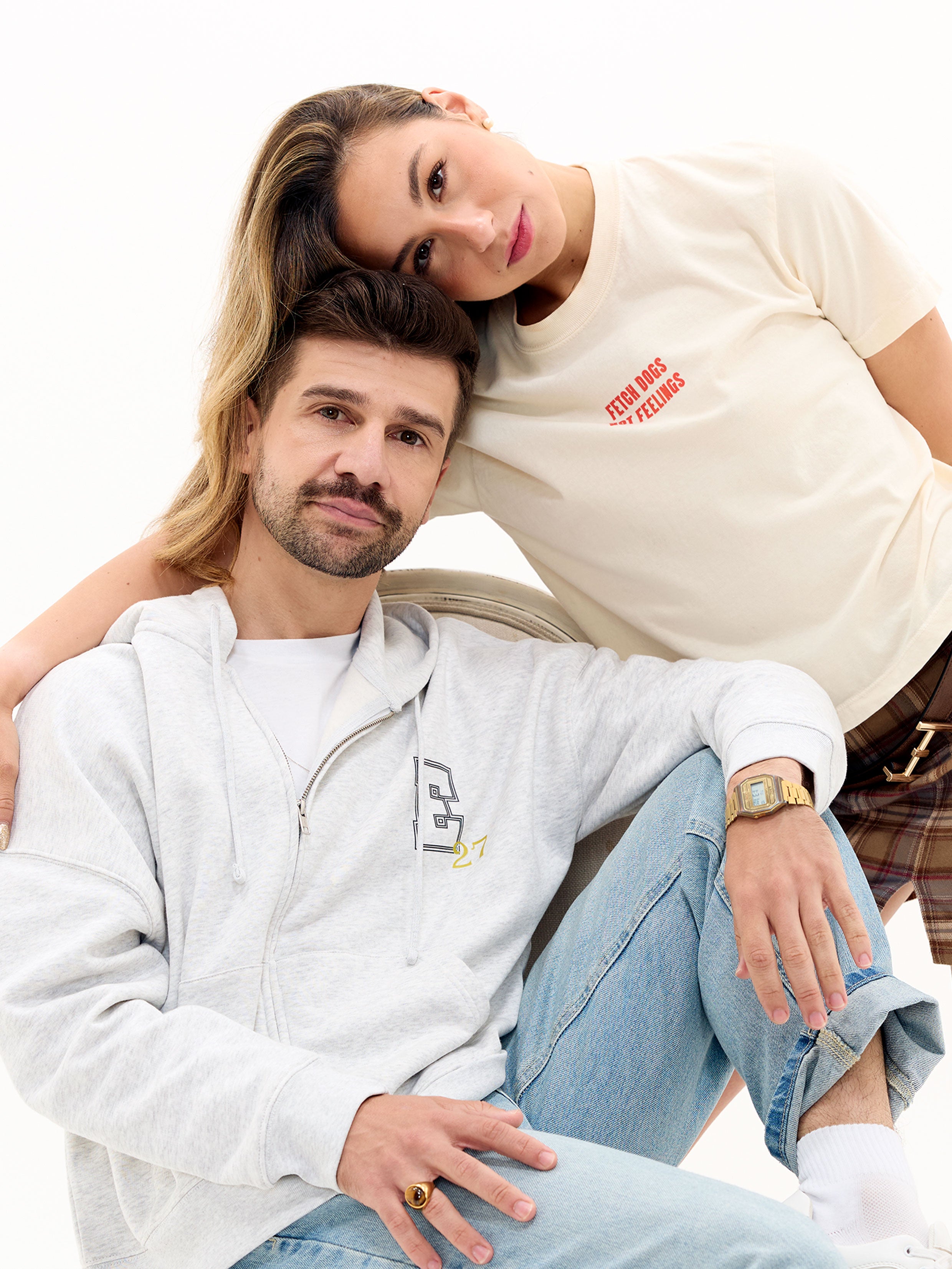Two people sitting close together, one wearing a grey zip hoodie with an E27 logo and the other in an off-white shirt with Fetch Dogs Not Feelings graphic on the front.