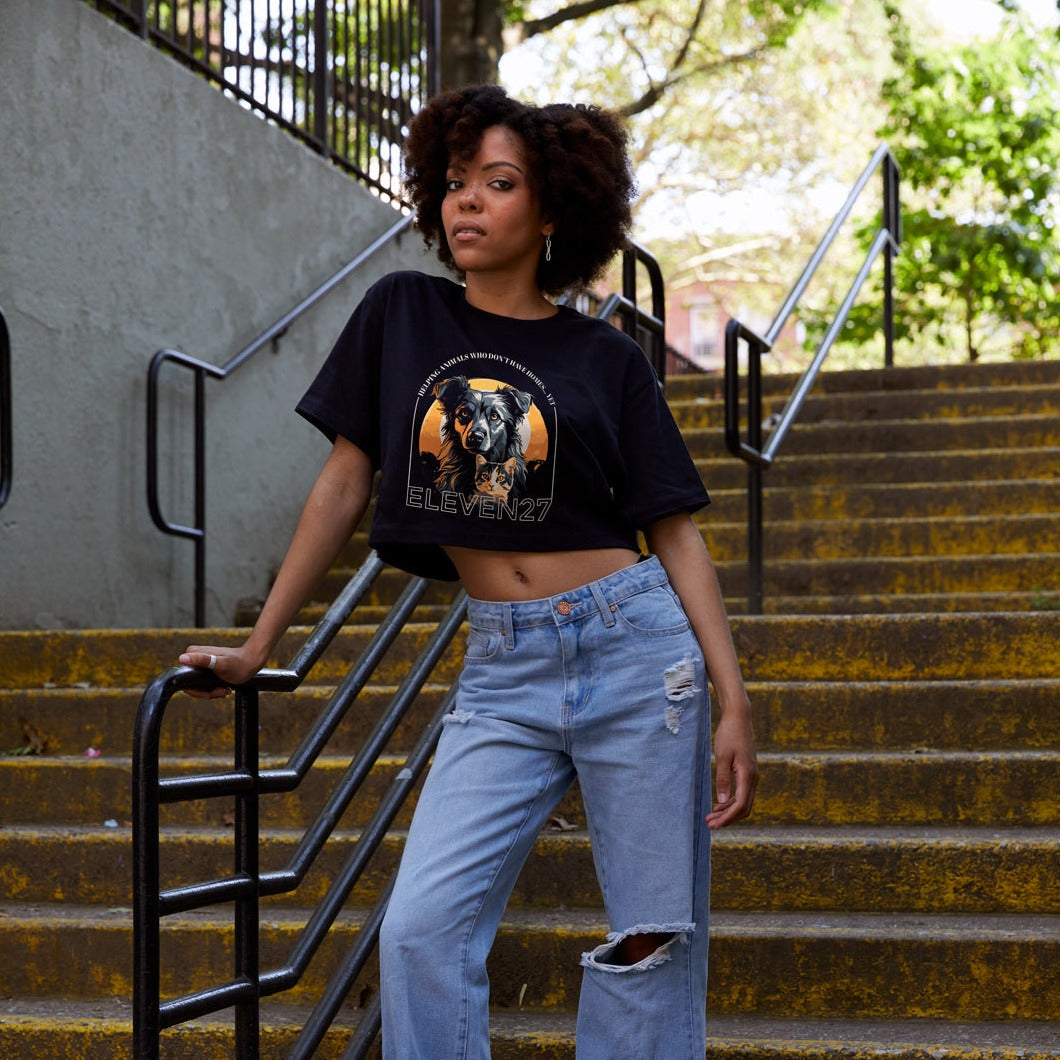 Lifestyle photo of a light skin model with natural curly hair on steps at a NYC park wearing Black cropped tshirt with graphic of a dog and a cat in a gray, orange, black color scheme. Tshirt sales support an animal shelter in need of support. Graphic also includes the brand name Eleven27 in ivory outline, with the words "Helping animals who don't have homes... yet". Model is wearing wide leg destructed jeans and Panda Nike sneakers