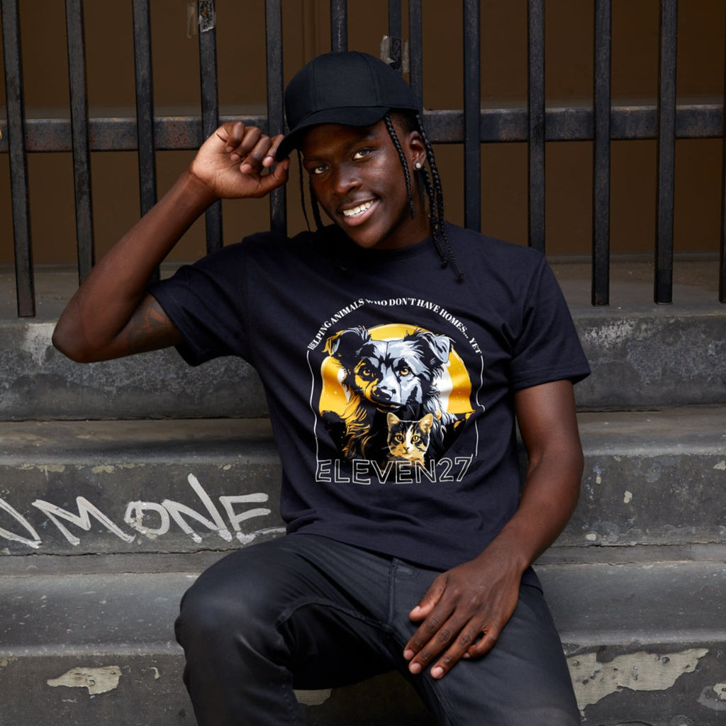 Afrian American male model wearing Black tshirt with graphic of a dog and a cat in a gray, orange, black color scheme. Tshirt sales support an animal shelter in need of support. Graphic also includes the brand name Eleven27 in ivory outline, with the words "Helping animals who don't have homes... yet". Model is sitting on a NYC stoop.