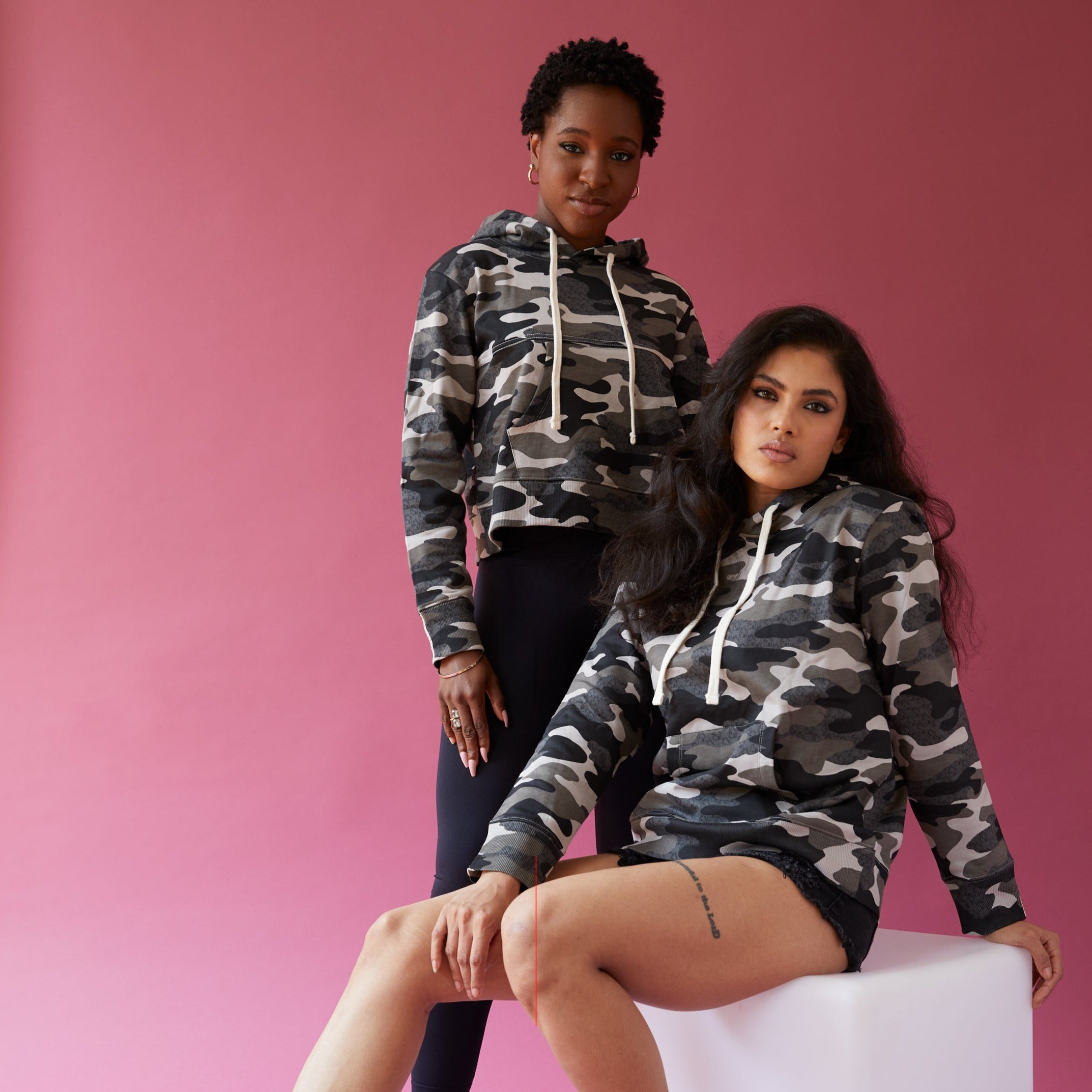 Photo of two models- one model is African American standing  in Shades of grey Camo printed cropped hoodie and black leggings. And an exotic model seated wearing the full length camo hoodie and black jean shorts