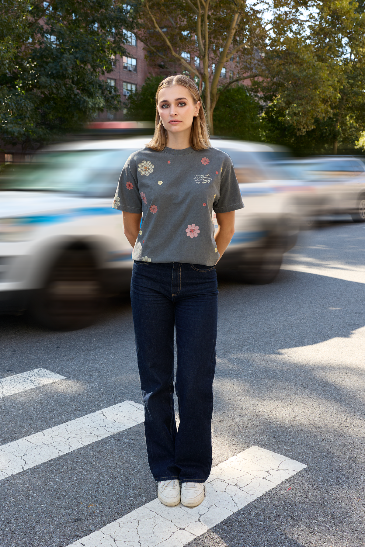 A woman stands on a city crosswalk wearing a gray Eleven27 floral graphic t-shirt with colorful floral prints, dark blue jeans, and white sneakers, with blurred cars and trees in the background.