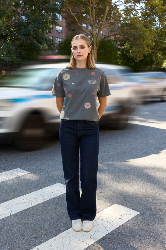 A woman stands on a city crosswalk wearing a gray Eleven27 floral graphic t-shirt with colorful floral prints, dark blue jeans, and white sneakers, with blurred cars and trees in the background.