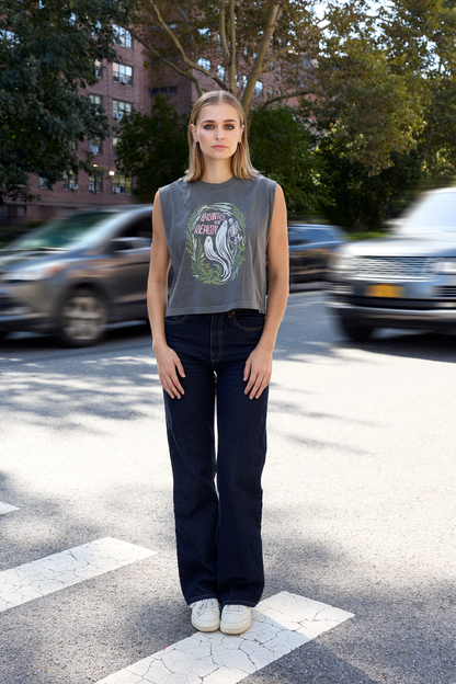 A woman stands on a city crosswalk wearing a gray sleeveless graphic T-shirt with a green and pink ghost design that says "Haunted by Reality", dark wide-leg jeans, and white sneakers. Cars and trees are blurred in the background