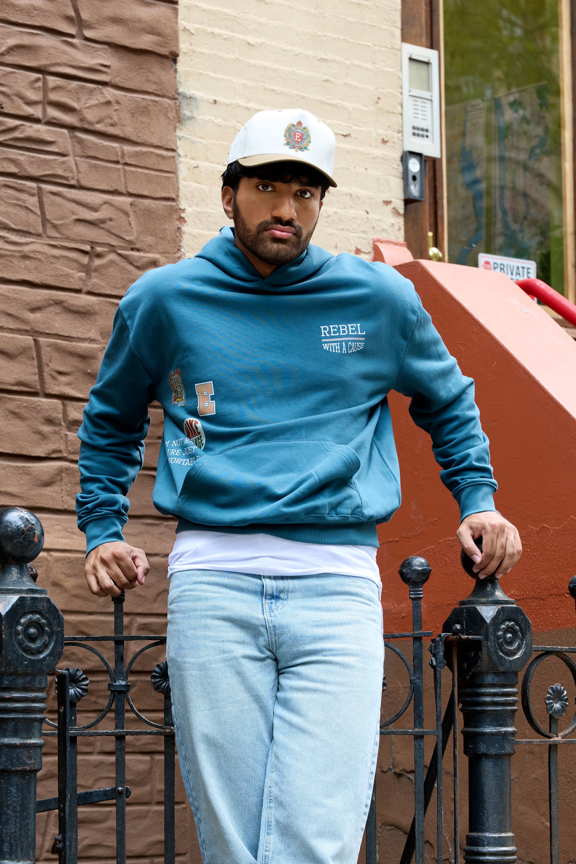 Man wearing a teal Eleven27 sweatshirt with 'Rebel With A Cause' text and light blue jeans standing against a brick wall.