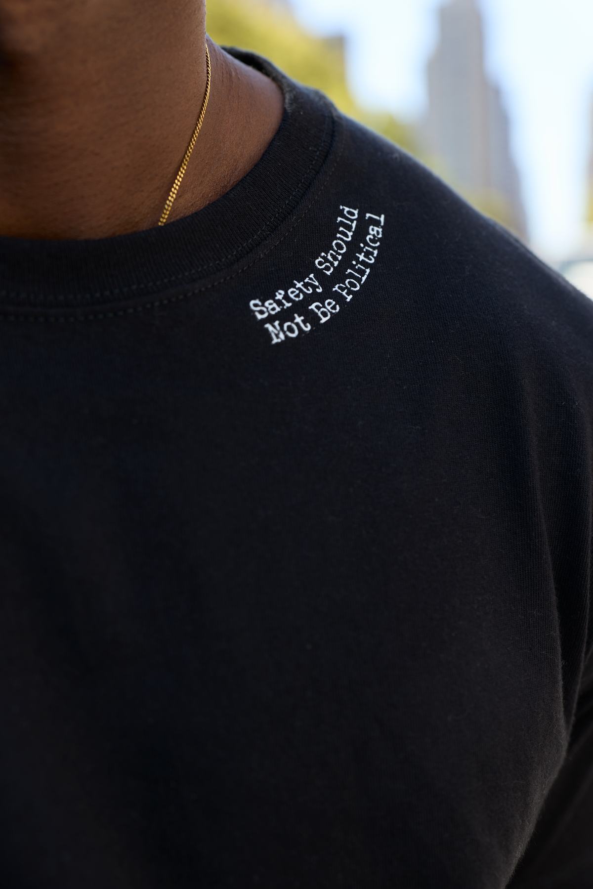 Close-up of a person wearing a black crewneck sweatshirt with white embroidered text reading 'Safety should Not Be political' on the left chest, along with a gold chain necklace.