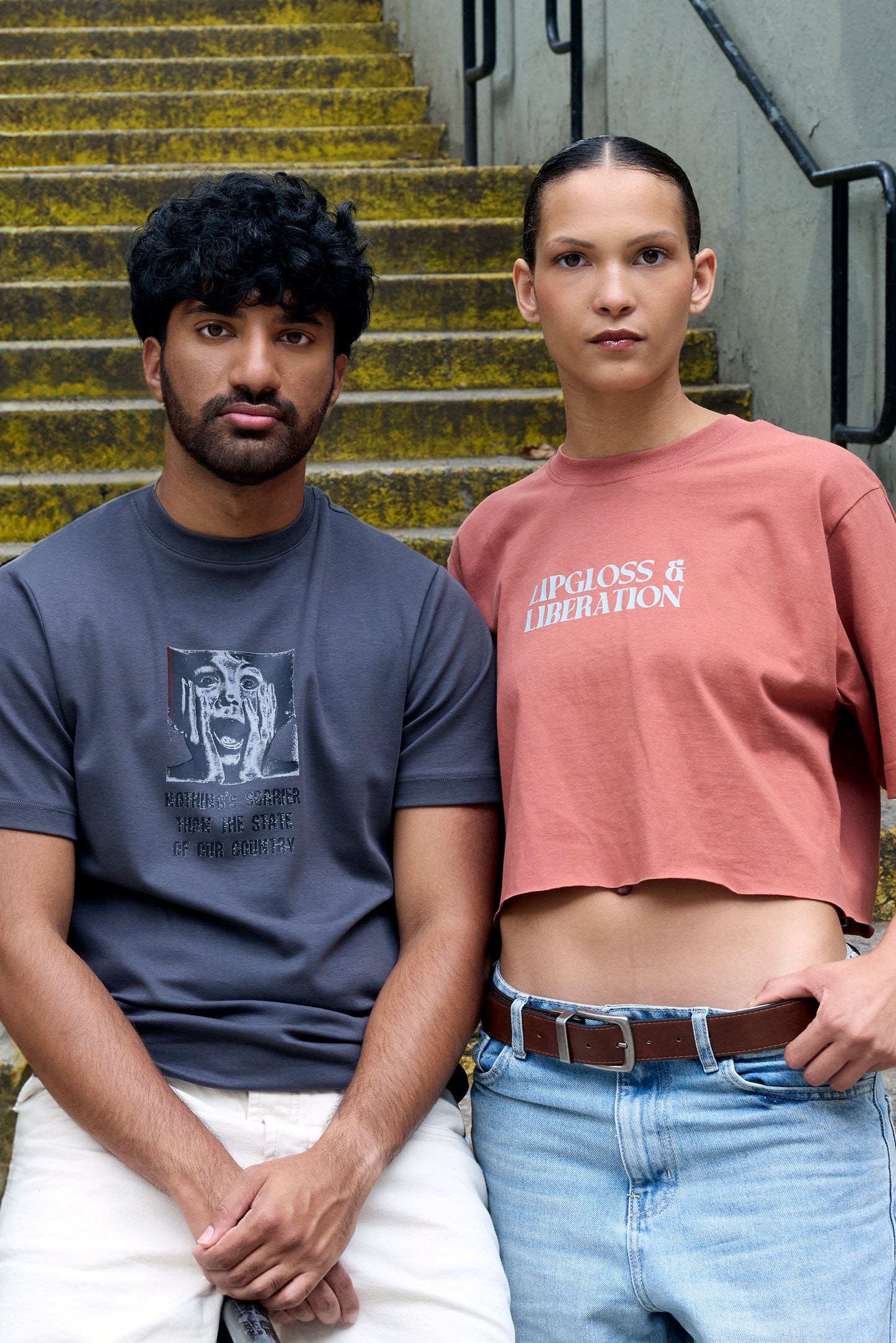 Two people standing on a set of outdoor stairs, one wearing a dark blue t-shirt and white pants, the other wearing a orange cropped Lipgloss & Liberation top and jeans.