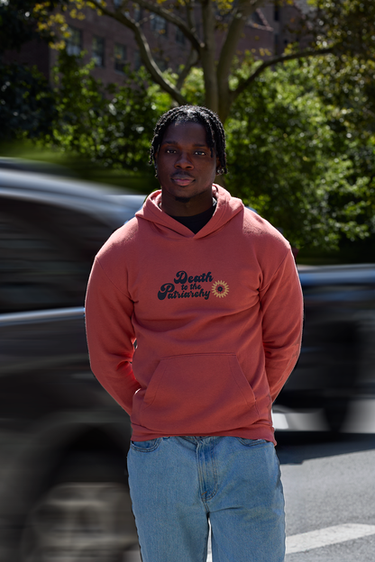 A man wearing a orange hoodie with 'Death To The Patriarchy' and a graphic printed on the chest, paired with light blue jeans, standing outdoors with a blurred car and trees in the background.