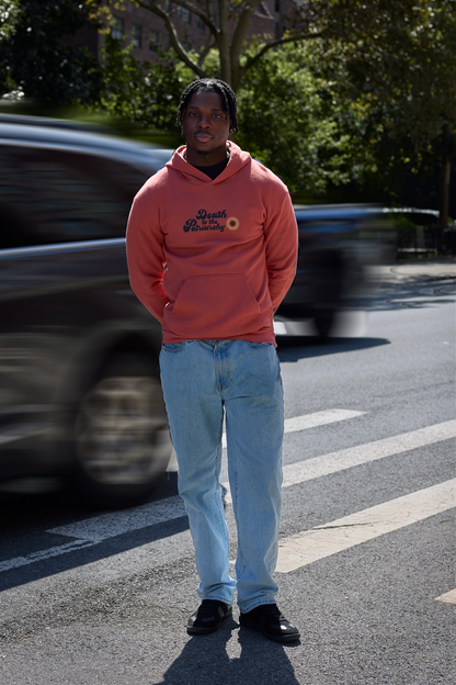 A man standing on a city street crosswalk wearing a orange hoodie with a 'Death To The Patriarchy' graphic print on the front and light blue jeans, with blurred cars in the background.