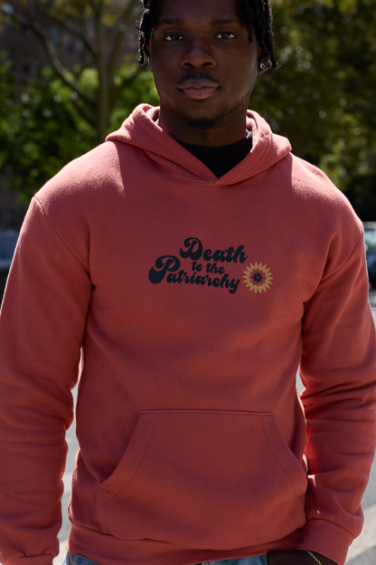 A man wearing a coral pink hoodie with the text 'Death to the Patriarchy' and a small sunflower graphic on the chest, photographed outdoors with trees in the background.