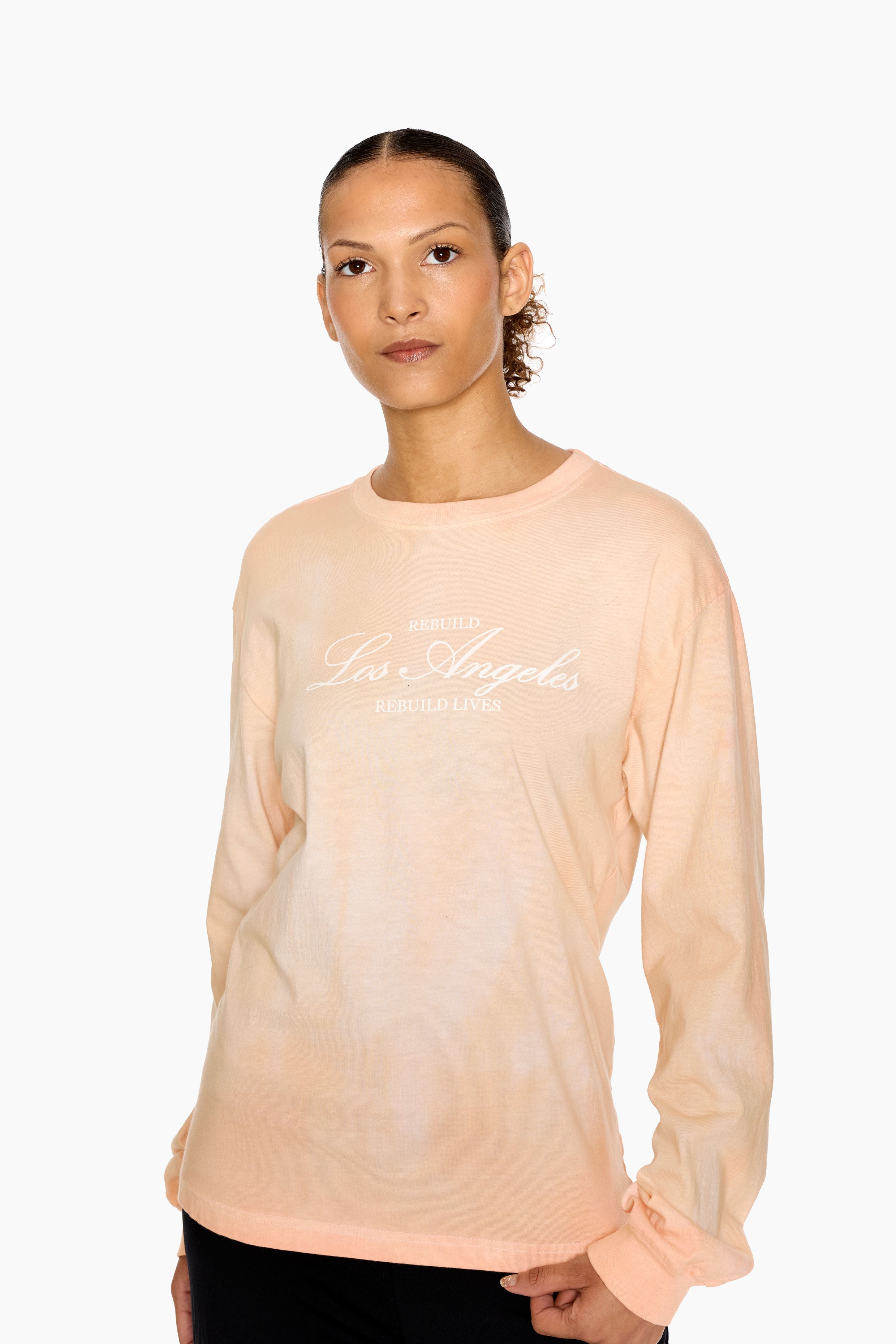 Woman wearing a peach long-sleeve top with text on a white background. Eleven27 long sleeve t-shirt, hand-dyed for a sunkissed look. "Rebuild Los Angeles, Rebuild Lives" graphic on the front.