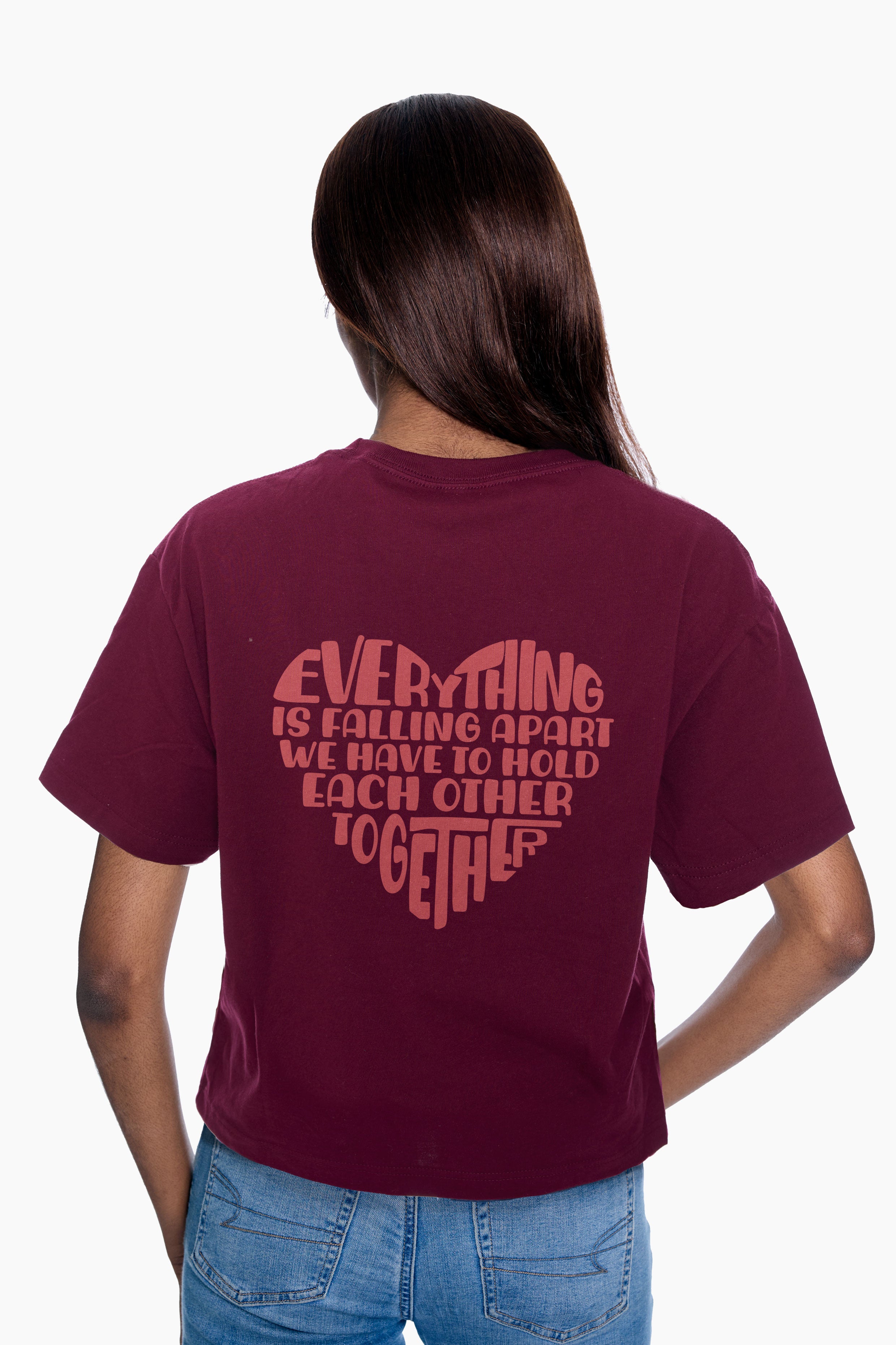 Person wearing a burgundy t-shirt with a "Everything is falling apart, we have to hold each other together" heart graphic on the back, standing against a white background.